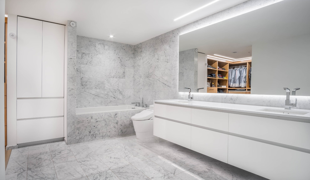 Modern bathroom with flat-panel cabinets, white cabinets, an alcove bath, a one-piece toilet, grey tiles, marble tiles, grey walls, a submerged sink, grey floors and white worktops.