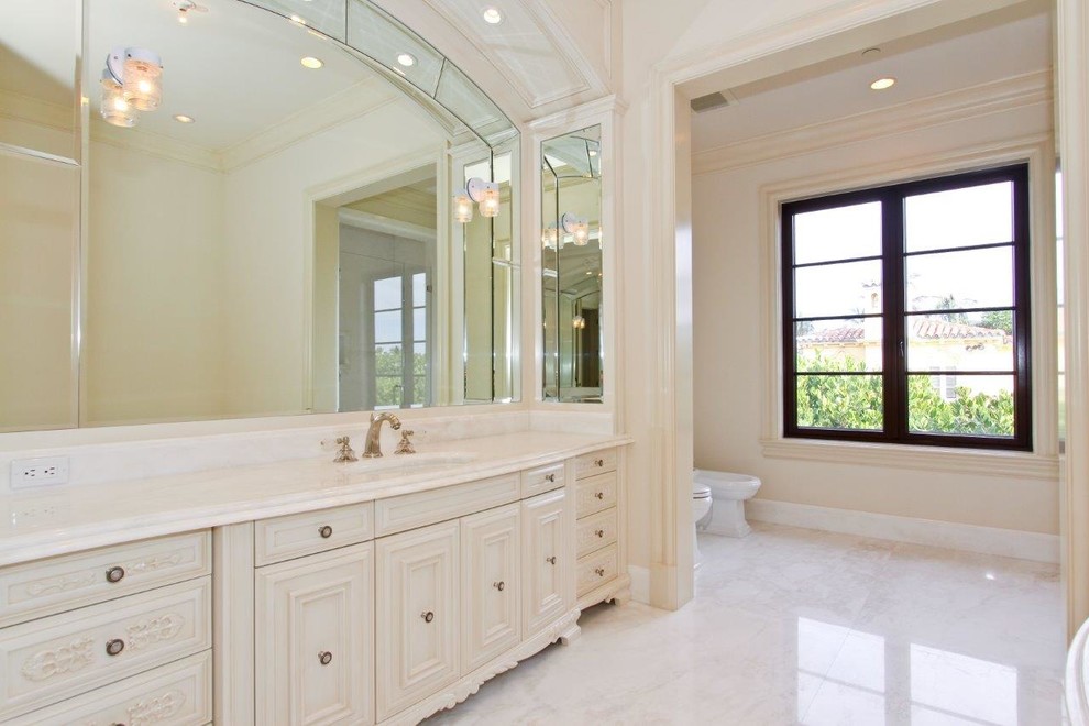 Cette image montre une grande salle de bain principale traditionnelle avec un placard en trompe-l'oeil, des portes de placard beiges, un bidet, un mur beige, un sol en marbre, un lavabo encastré, un plan de toilette en marbre et un sol blanc.