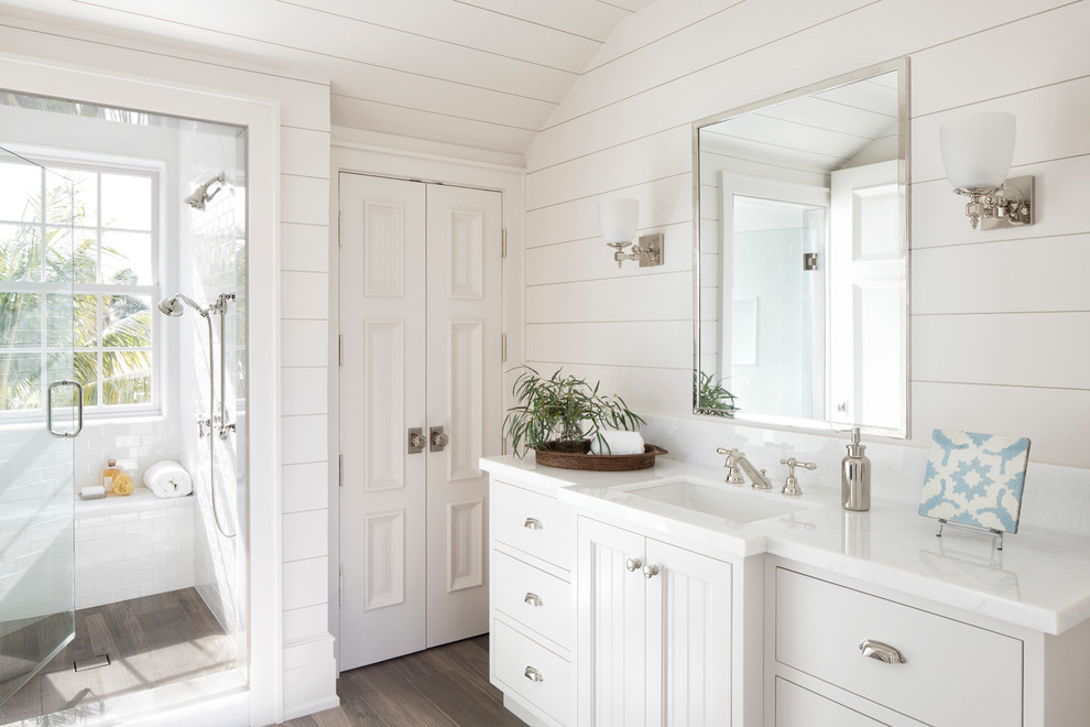 Cette image montre une salle de bain marine avec des portes de placard blanches, un carrelage blanc, un mur blanc, un lavabo encastré, un sol marron, une cabine de douche à porte battante, un plan de toilette blanc et une fenêtre.