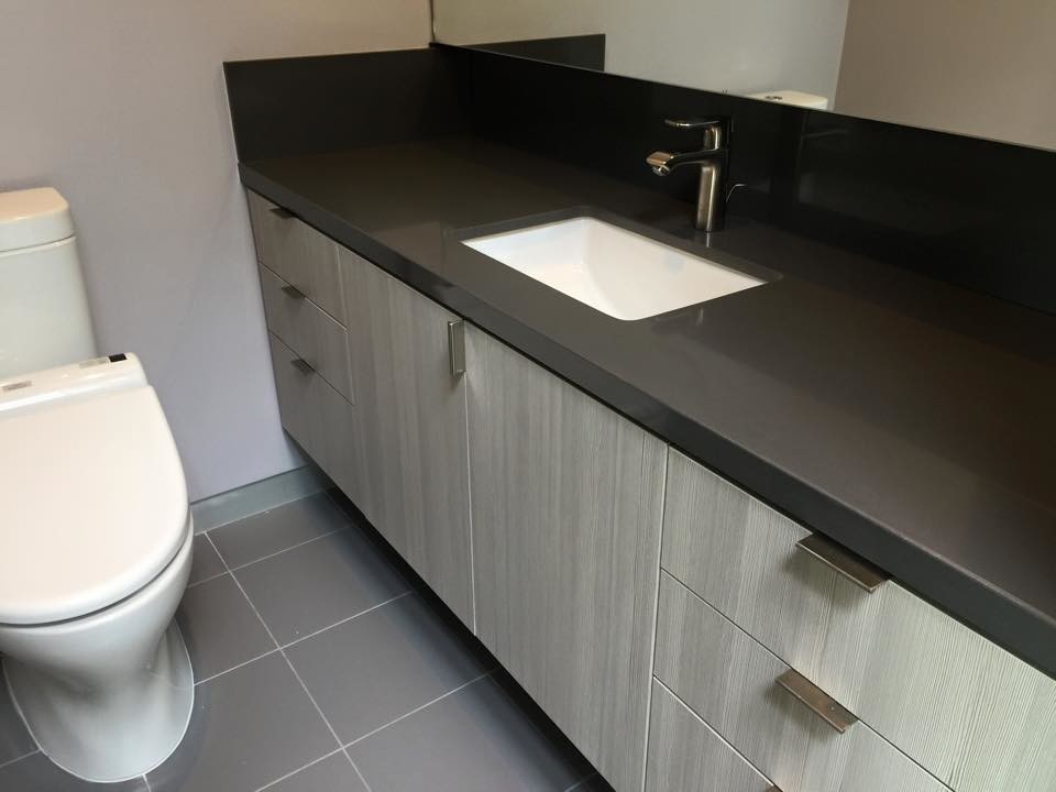 Cette image montre une salle de bain minimaliste de taille moyenne avec un placard à porte plane, des portes de placard grises, WC à poser, un mur gris, un sol en carrelage de porcelaine, un lavabo encastré, un plan de toilette en surface solide, un sol gris et un plan de toilette gris.