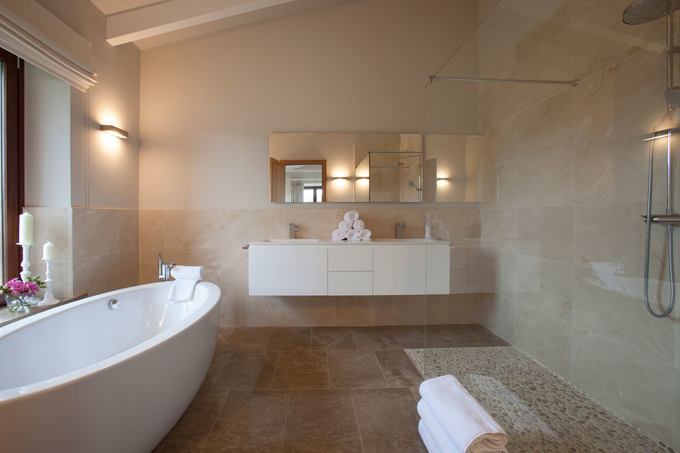 Coastal bathroom in Palma de Mallorca with a freestanding bath, beige tiles, beige walls, an integrated sink, flat-panel cabinets, white cabinets, a corner shower, pebble tile flooring, beige floors and white worktops.