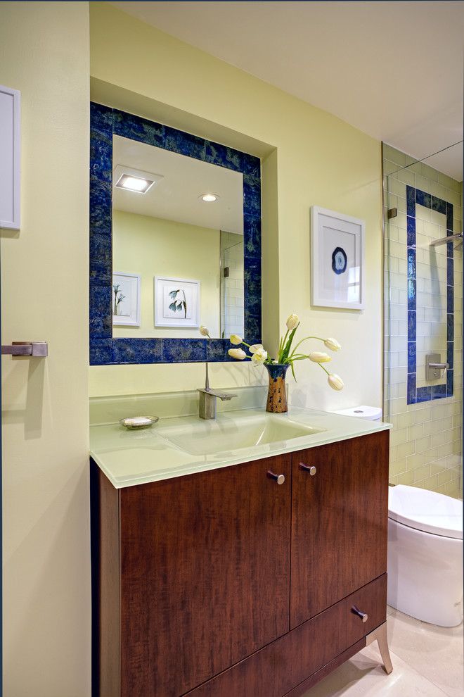 Cette photo montre une petite salle d'eau tendance en bois foncé avec un placard à porte plane, un plan de toilette en verre, un lavabo intégré, WC à poser, un mur vert, un sol en calcaire, un carrelage vert, un carrelage en pâte de verre et un sol beige.