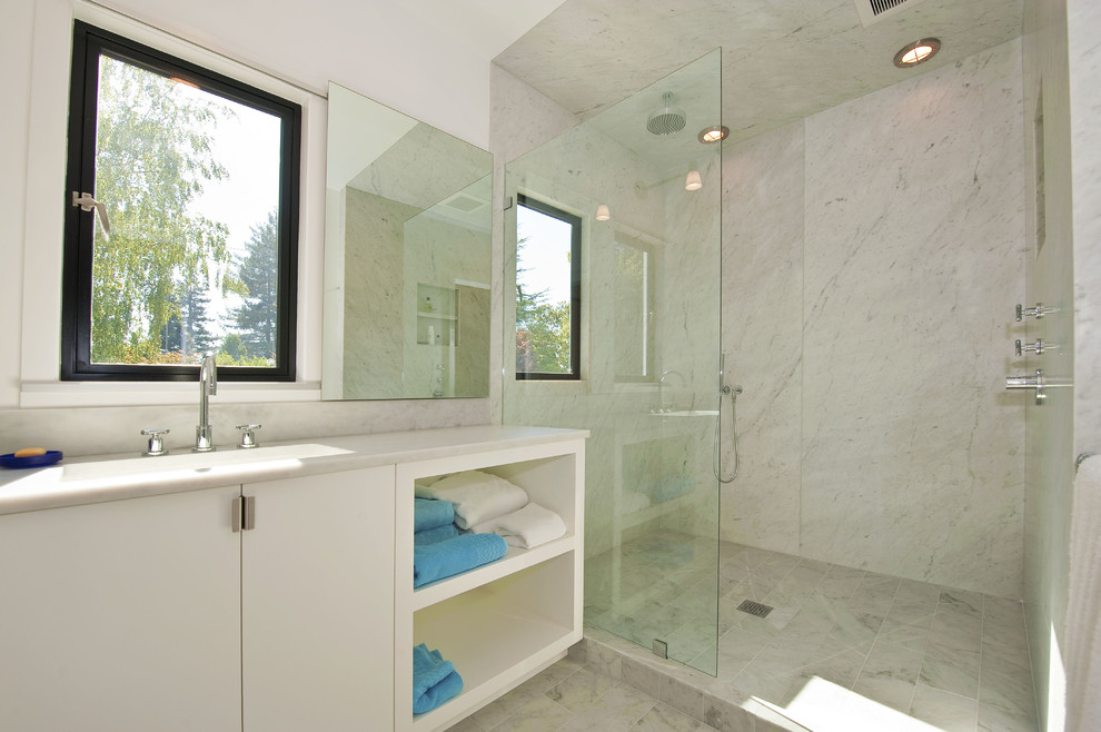 Inredning av ett modernt badrum, med ett integrerad handfat, släta luckor, vita skåp, en öppen dusch, vit kakel och med dusch som är öppen