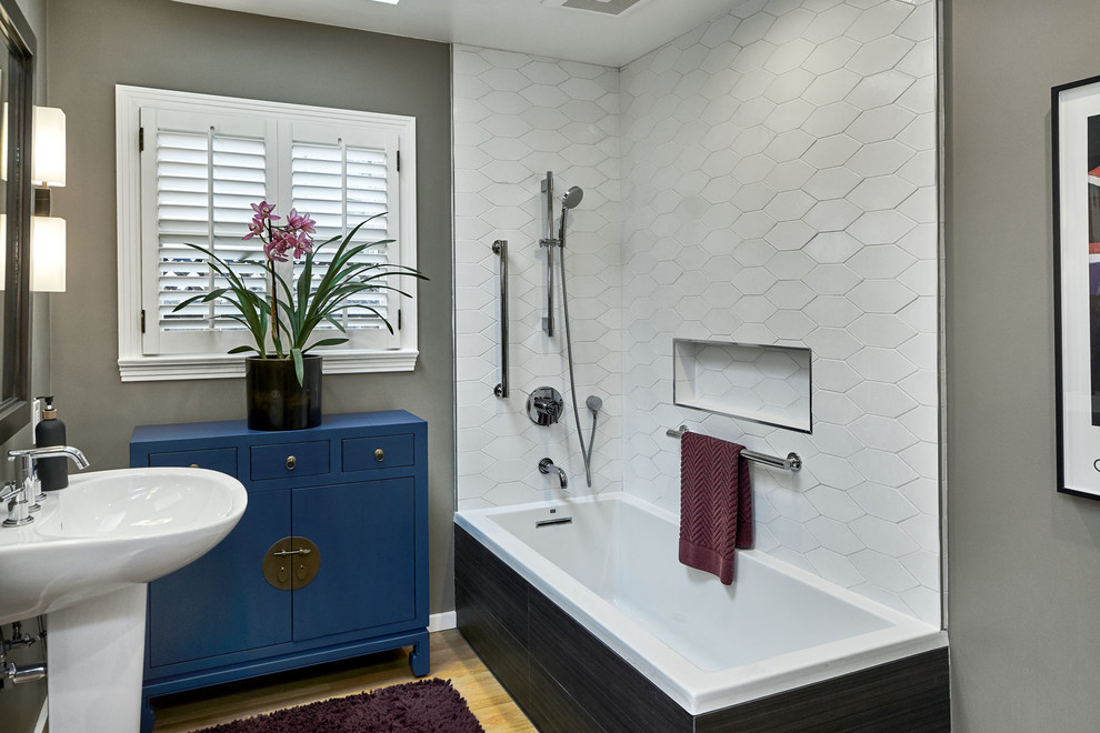 Bild på ett litet vintage vit vitt badrum med dusch, med släta luckor, blå skåp, ett hörnbadkar, en dusch/badkar-kombination, en toalettstol med hel cisternkåpa, vit kakel, keramikplattor, grå väggar, ljust trägolv, ett piedestal handfat, gult golv och med dusch som är öppen