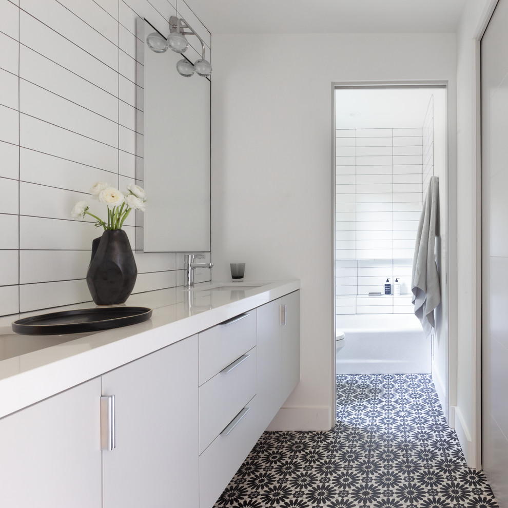 На фото: ванная комната в современном стиле с плоскими фасадами, серыми фасадами, ванной в нише, душем над ванной, белой плиткой, белыми стенами, полом из цементной плитки, врезной раковиной, разноцветным полом и белой столешницей