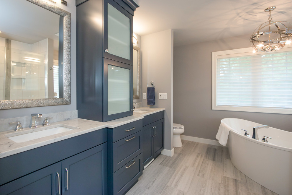 This is an example of a medium sized classic ensuite bathroom in Cedar Rapids with recessed-panel cabinets, blue cabinets, a freestanding bath, grey walls, porcelain flooring, engineered stone worktops, grey floors and white worktops.