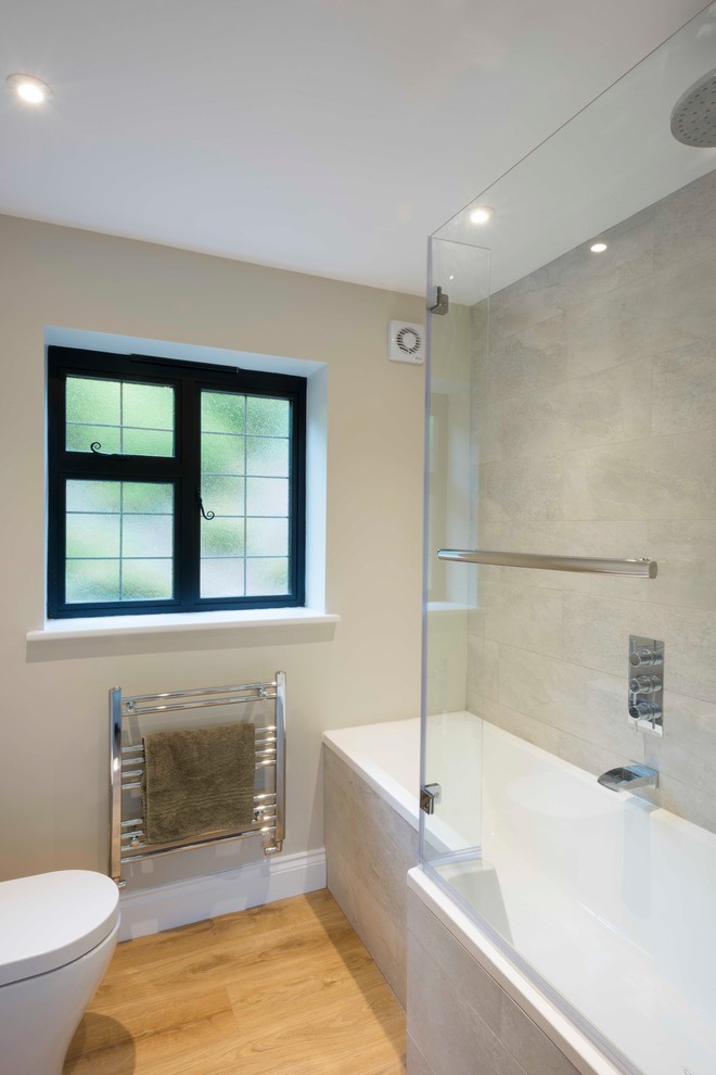 Photo of a large country family bathroom in Sussex with flat-panel cabinets, white cabinets, a built-in bath, a shower/bath combination, a wall mounted toilet, grey tiles, limestone tiles, white walls, medium hardwood flooring, a console sink, brown floors and a hinged door.