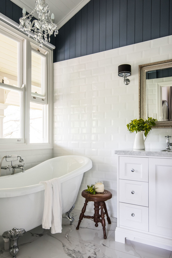 Diseño de cuarto de baño principal de estilo de casa de campo de tamaño medio con puertas de armario blancas, bañera con patas, baldosas y/o azulejos blancos, baldosas y/o azulejos de cemento, paredes azules, suelo de baldosas de porcelana, lavabo encastrado, encimera de mármol y armarios con paneles empotrados