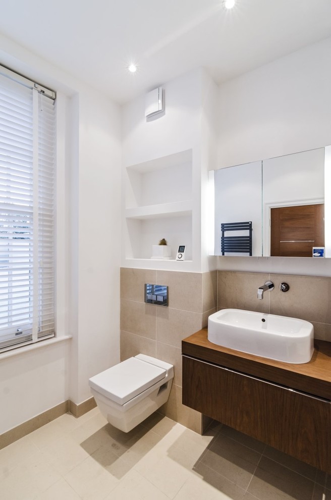 Photo of a modern bathroom in London with a vessel sink, flat-panel cabinets, dark wood cabinets, wooden worktops, a wall mounted toilet and beige tiles.