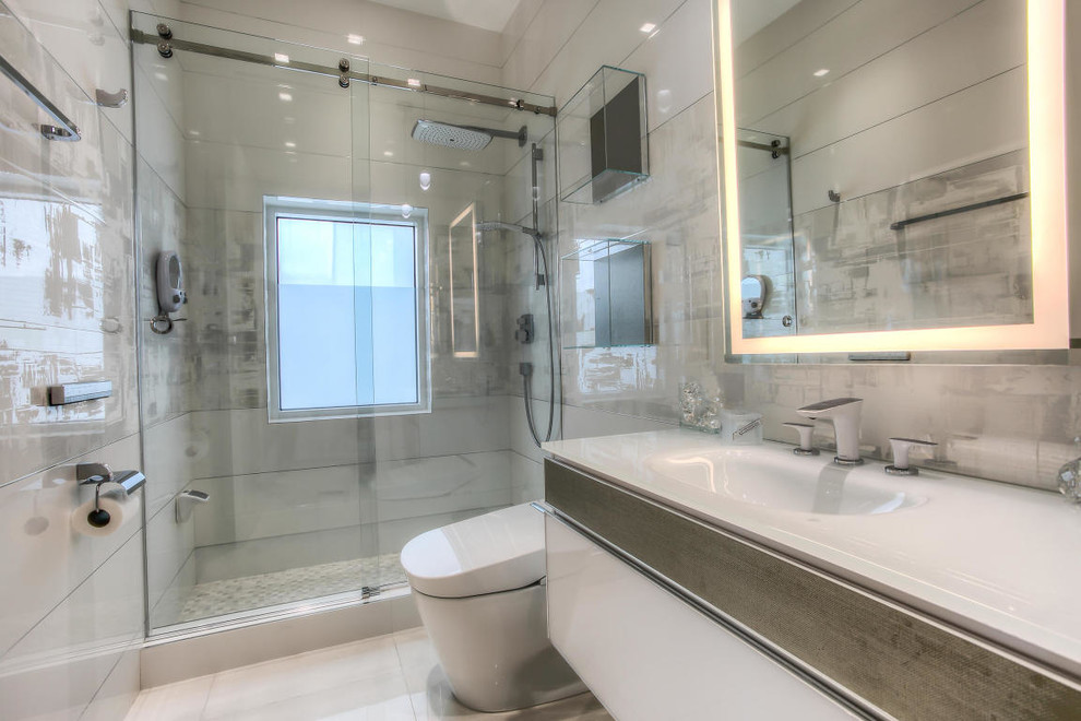 Cette image montre une salle de bain design de taille moyenne avec un placard à porte plane, des portes de placard blanches, un bidet, un carrelage gris, des carreaux de porcelaine, un mur gris, un sol en carrelage de porcelaine, un lavabo intégré et un plan de toilette en verre.