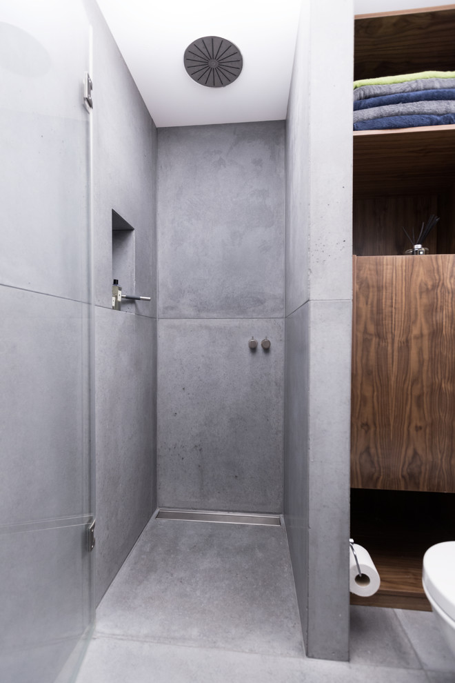 Modelo de cuarto de baño actual de tamaño medio con puertas de armario de madera en tonos medios, encimera de madera, ducha abierta, sanitario de pared, baldosas y/o azulejos grises, suelo de cemento y aseo y ducha