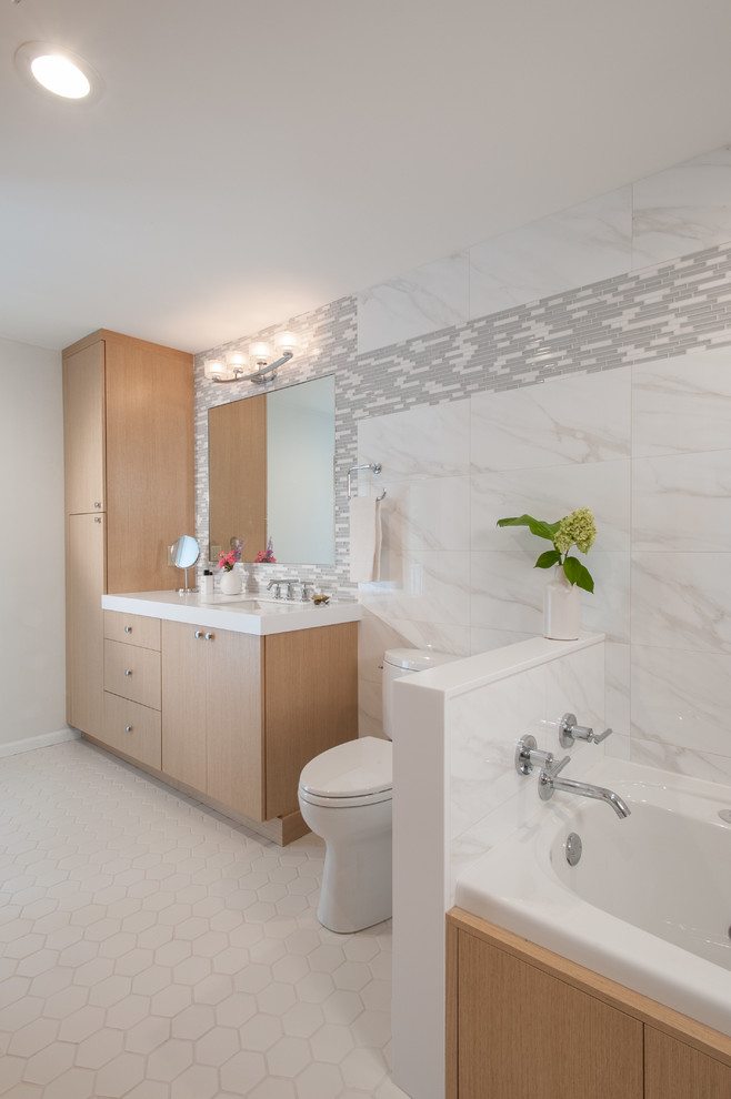 Inredning av ett modernt badrum, med släta luckor, skåp i ljust trä, en toalettstol med separat cisternkåpa, vit kakel, stenkakel och ett platsbyggt badkar