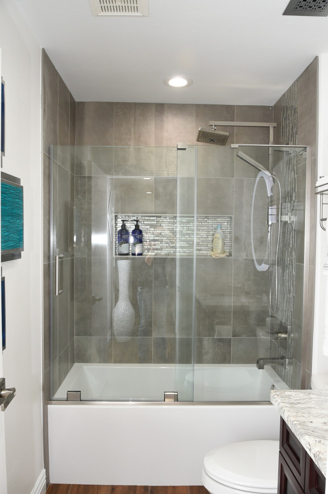 Photo of a medium sized modern family bathroom in Phoenix with recessed-panel cabinets, dark wood cabinets, an alcove bath, a shower/bath combination, a one-piece toilet, grey tiles, porcelain tiles, white walls, porcelain flooring, a submerged sink, granite worktops, multi-coloured floors, a hinged door, grey worktops, a wall niche, a single sink and a built in vanity unit.