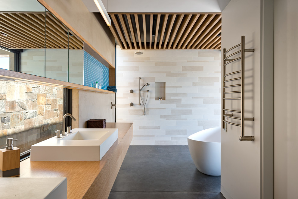 Modern inredning av ett brun brunt badrum, med släta luckor, skåp i mellenmörkt trä, ett fristående badkar, beige kakel, beige väggar, betonggolv, ett fristående handfat, träbänkskiva och grått golv