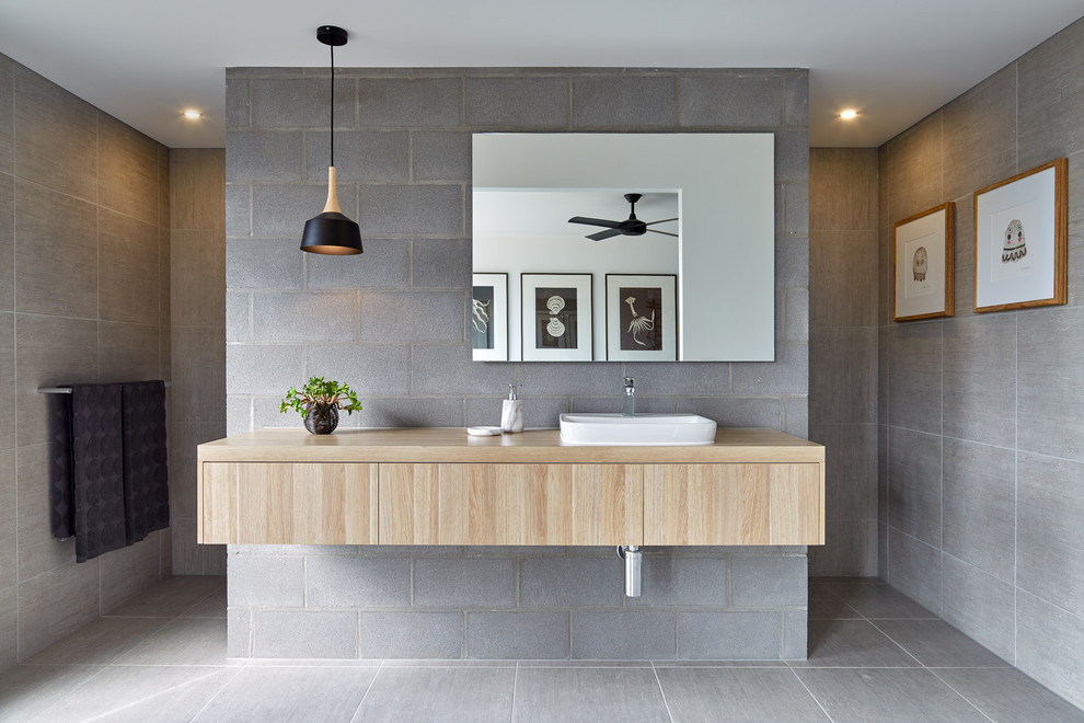 Modelo de cuarto de baño rectangular moderno con armarios con paneles lisos, puertas de armario de madera clara, baldosas y/o azulejos grises, lavabo sobreencimera, encimera de madera y ducha abierta