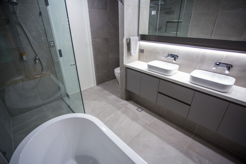 Design ideas for a medium sized modern ensuite bathroom in Brisbane with flat-panel cabinets, grey cabinets, a freestanding bath, a corner shower, a two-piece toilet, grey tiles, porcelain tiles, grey walls, porcelain flooring, a vessel sink, engineered stone worktops, grey floors, a hinged door and white worktops.