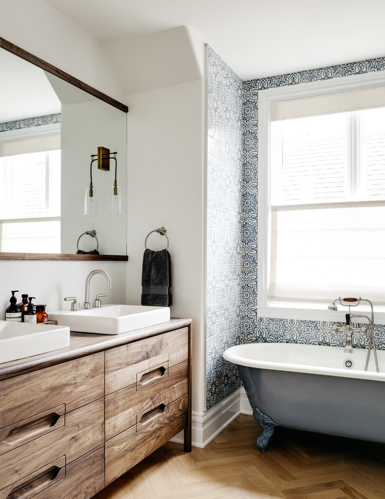 Пример оригинального дизайна: ванная комната в стиле неоклассика (современная классика) с темными деревянными фасадами, ванной на ножках, синей плиткой, белой плиткой, белыми стенами, паркетным полом среднего тона, накладной раковиной, столешницей из дерева, коричневым полом, коричневой столешницей, зеркалом с подсветкой и плоскими фасадами