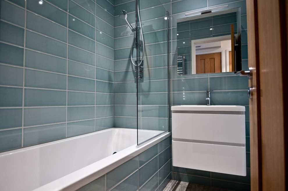 This is an example of a small modern family bathroom in London with flat-panel cabinets, white cabinets, a built-in bath, a shower/bath combination, a one-piece toilet, grey tiles, grey walls, a wall-mounted sink and ceramic tiles.