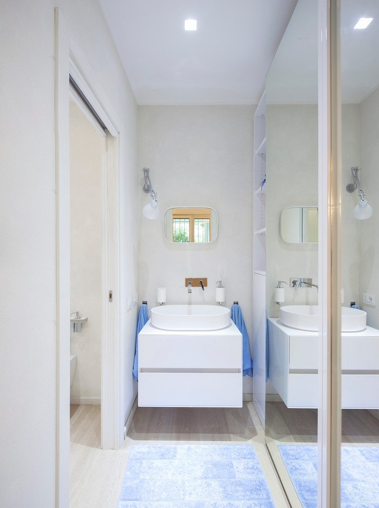 Diseño de cuarto de baño minimalista con armarios con paneles lisos, puertas de armario blancas, suelo de madera clara, lavabo sobreencimera y paredes blancas