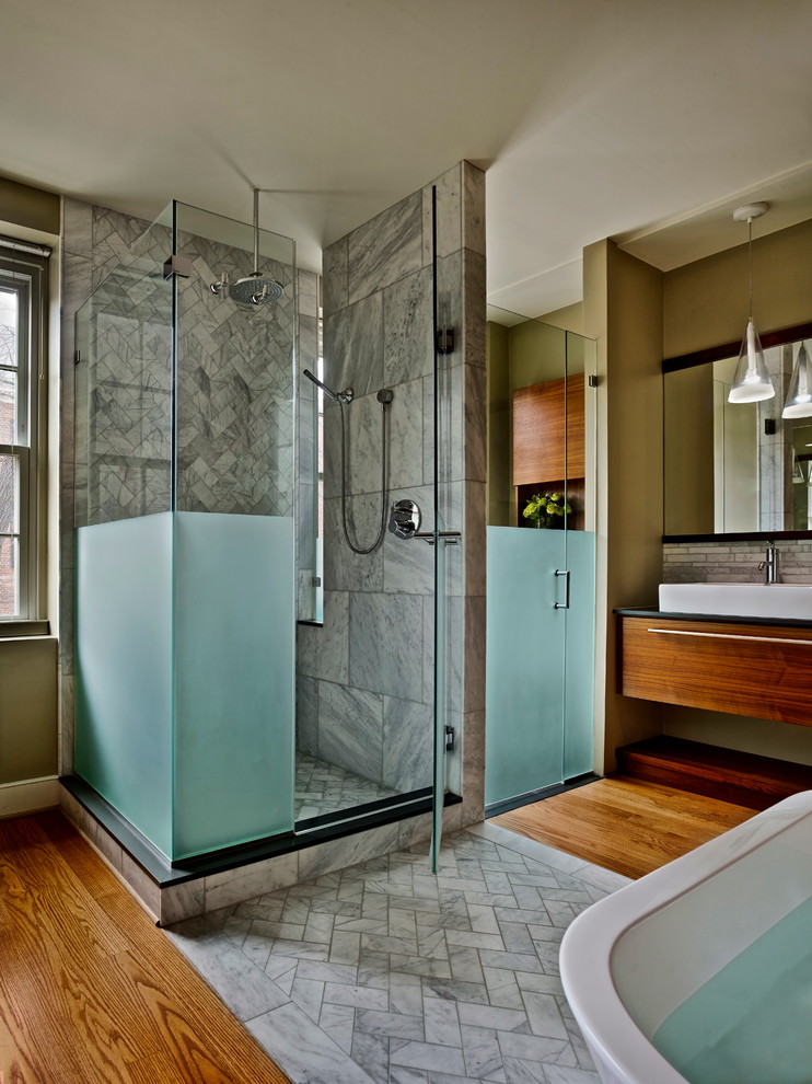 Modern inredning av ett mellanstort en-suite badrum, med släta luckor, bruna skåp, ett badkar med tassar, en dusch i en alkov, en toalettstol med separat cisternkåpa, vit kakel, marmorkakel, beige väggar, mellanmörkt trägolv, ett fristående handfat, marmorbänkskiva och dusch med gångjärnsdörr