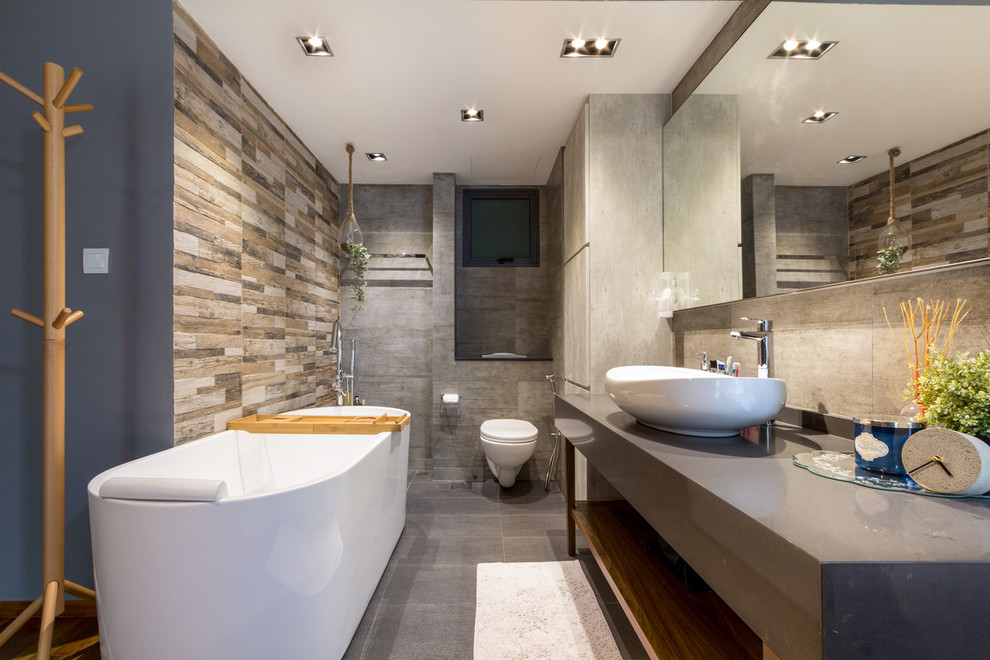 Cette image montre une salle de bain design avec un placard sans porte, une baignoire indépendante, WC suspendus, un carrelage gris, un mur gris, une vasque, un sol gris et un plan de toilette gris.