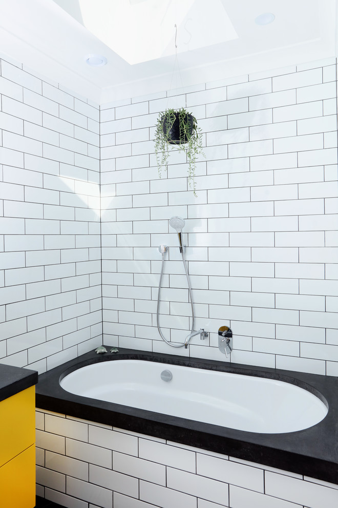 Пример оригинального дизайна: детская ванная комната среднего размера в современном стиле с полновстраиваемой ванной, плиткой кабанчик, полом из керамогранита, столешницей из бетона, плоскими фасадами, желтыми фасадами, белой плиткой, белыми стенами, врезной раковиной, черным полом, серой столешницей, тумбой под одну раковину, подвесной тумбой и кирпичными стенами