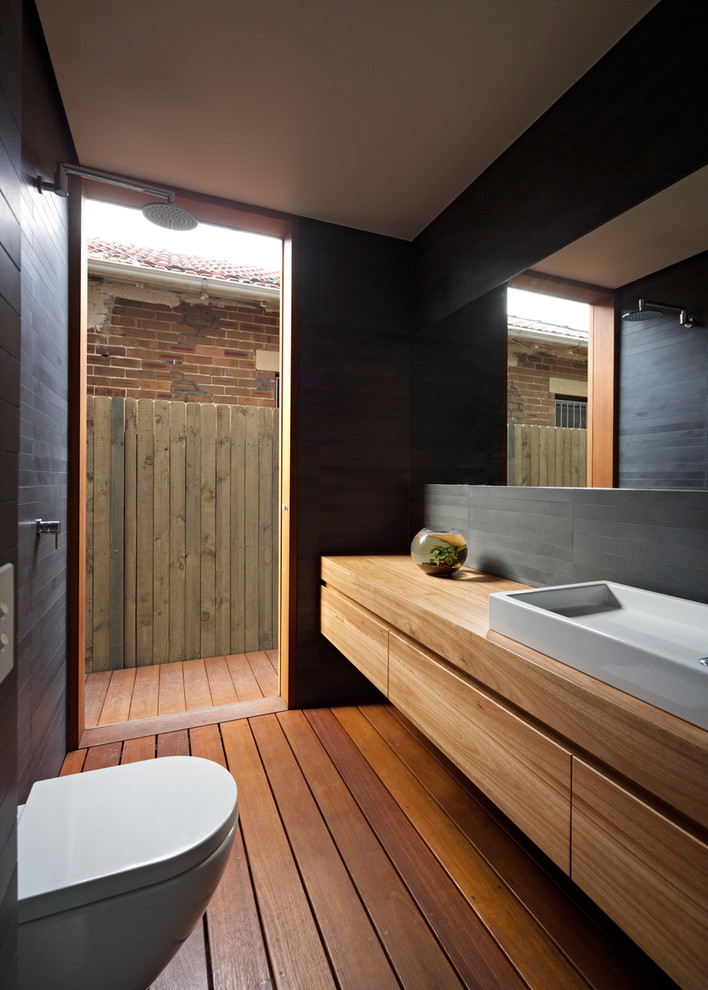 Foto på ett mellanstort funkis badrum för barn, med ett fristående handfat, släta luckor, skåp i mellenmörkt trä, träbänkskiva, en öppen dusch, en vägghängd toalettstol, grå kakel, stenkakel, grå väggar och mellanmörkt trägolv