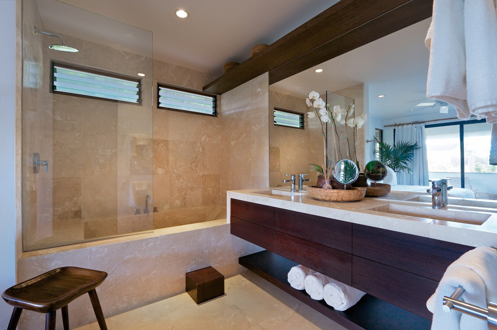 Medium sized world-inspired ensuite bathroom in Hawaii with flat-panel cabinets, dark wood cabinets, an alcove bath, a one-piece toilet, beige tiles, travertine tiles, beige walls, travertine flooring, an integrated sink, limestone worktops, beige floors, an open shower, a shower/bath combination and beige worktops.