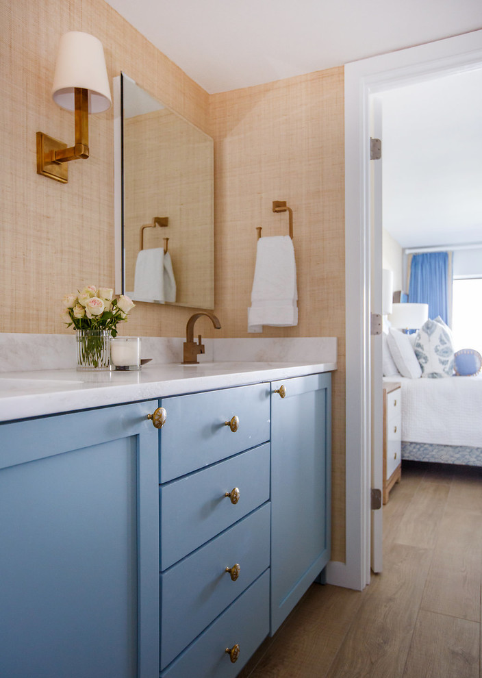 Modelo de cuarto de baño costero de tamaño medio con armarios estilo shaker, puertas de armario azules, paredes beige, suelo de madera en tonos medios, lavabo bajoencimera y encimera de acrílico