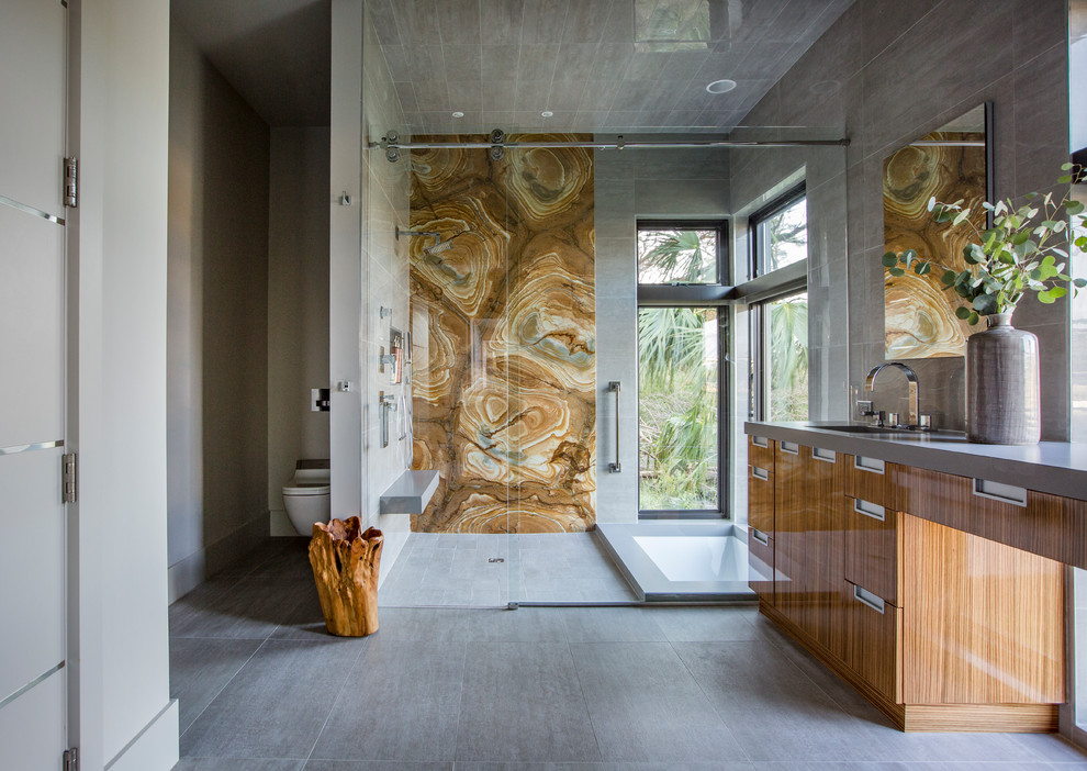 Modern inredning av ett stort grå grått en-suite badrum, med släta luckor, skåp i mellenmörkt trä, ett undermonterat badkar, en dusch i en alkov, en vägghängd toalettstol, brun kakel, stenhäll, grå väggar, grått golv och dusch med skjutdörr