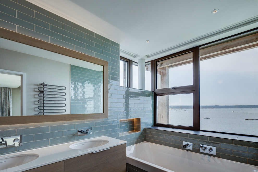 Modernes Badezimmer mit flächenbündigen Schrankfronten, dunklen Holzschränken, Einbaubadewanne, blauen Fliesen, Unterbauwaschbecken und weißer Waschtischplatte in Dorset
