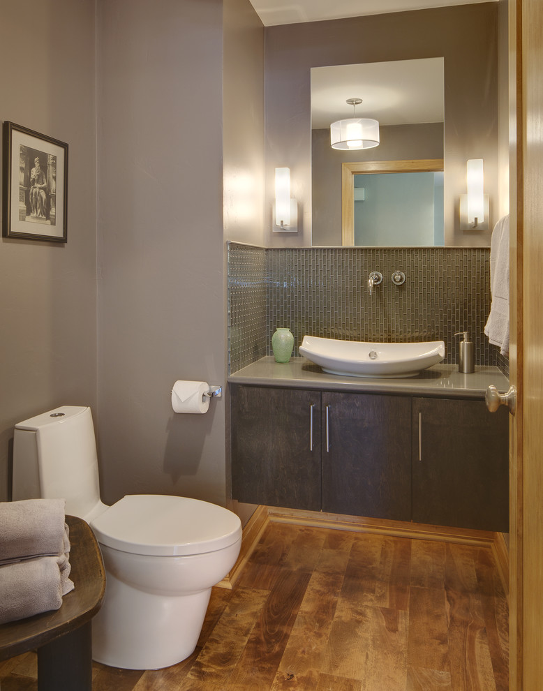 Inredning av ett retro litet badrum, med ett fristående handfat, släta luckor, grå skåp, bänkskiva i kvarts, en toalettstol med hel cisternkåpa, grå kakel, glaskakel, grå väggar och mellanmörkt trägolv