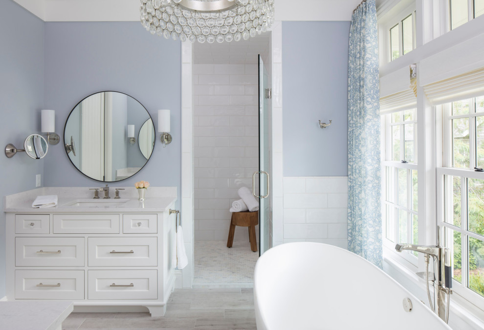 Пример оригинального дизайна: большая главная ванная комната в стиле неоклассика (современная классика) с фасадами с утопленной филенкой, белыми фасадами, отдельно стоящей ванной, душевой комнатой, унитазом-моноблоком, белой плиткой, синими стенами, полом из плитки под дерево, врезной раковиной, столешницей из искусственного кварца, серым полом, душем с распашными дверями и белой столешницей