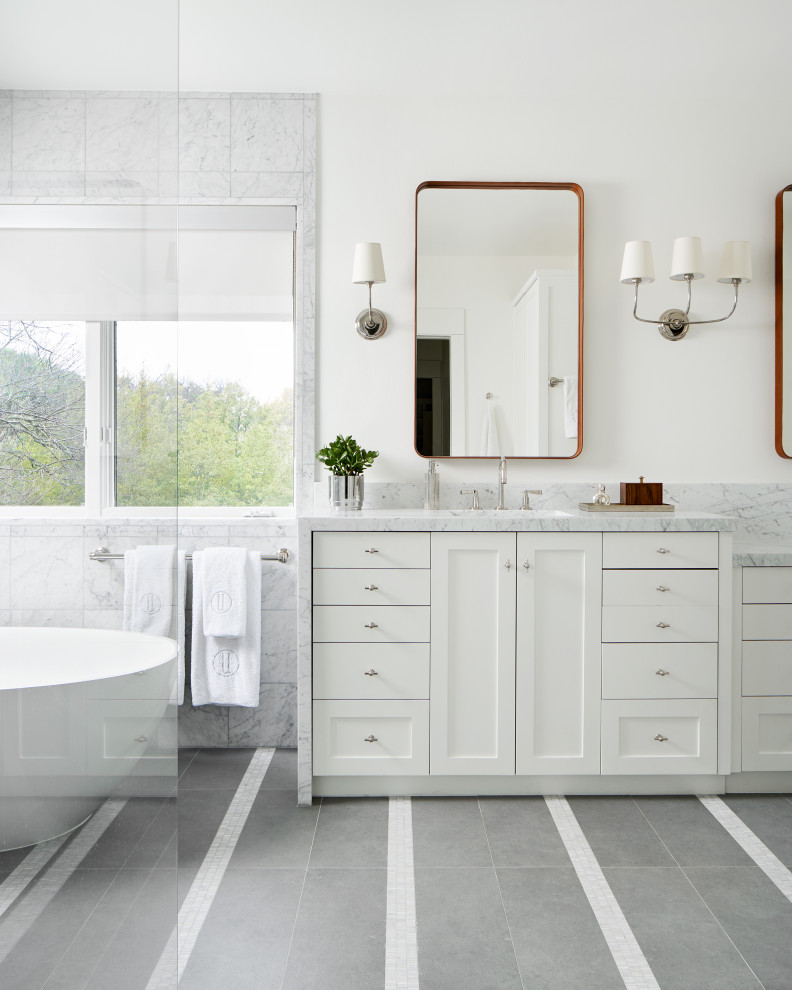 Klassisk inredning av ett stort vit vitt badrum, med möbel-liknande, vita skåp, ett fristående badkar, en dubbeldusch, en toalettstol med hel cisternkåpa, vit kakel, marmorkakel, vita väggar, klinkergolv i porslin, ett undermonterad handfat, marmorbänkskiva och med dusch som är öppen
