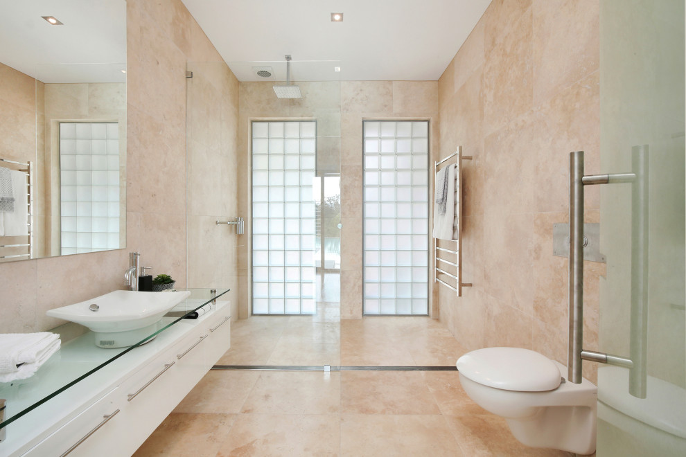 Bild på ett badrum, med släta luckor, vita skåp, en kantlös dusch, beige kakel, ett fristående handfat, bänkskiva i glas, beiget golv och med dusch som är öppen