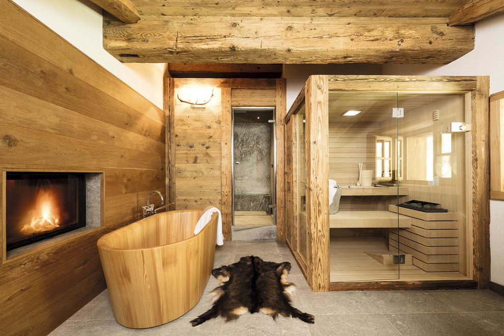 Cette photo montre un grand sauna montagne avec une baignoire indépendante, un espace douche bain, un mur marron, un sol en calcaire, un sol gris et une cabine de douche à porte battante.