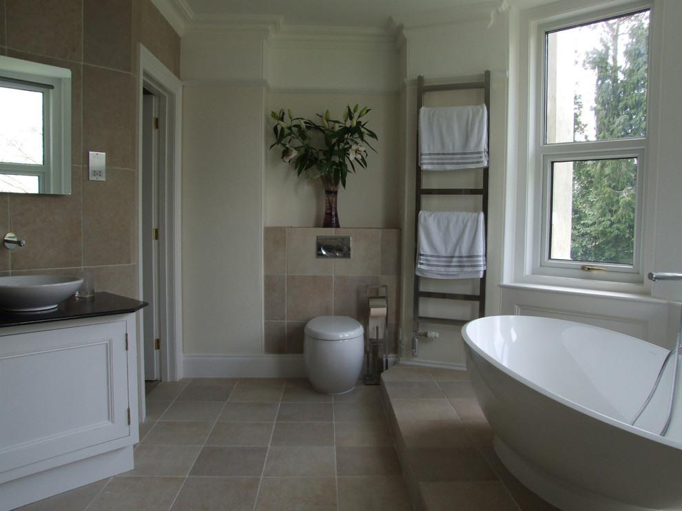 This is an example of an expansive contemporary ensuite bathroom in Cornwall with a vessel sink, freestanding cabinets, white cabinets, granite worktops, a freestanding bath, an alcove shower, a wall mounted toilet, beige tiles, stone tiles, beige walls and travertine flooring.