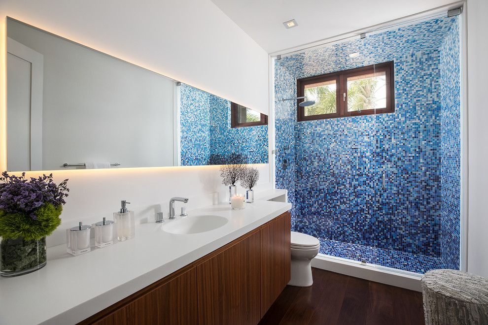 Mittelgroßes Modernes Badezimmer mit flächenbündigen Schrankfronten, blauen Fliesen, weißer Wandfarbe, dunklem Holzboden, integriertem Waschbecken, Quarzwerkstein-Waschtisch, braunem Boden, weißer Waschtischplatte, dunklen Holzschränken, Duschnische, Mosaikfliesen und Falttür-Duschabtrennung in Miami