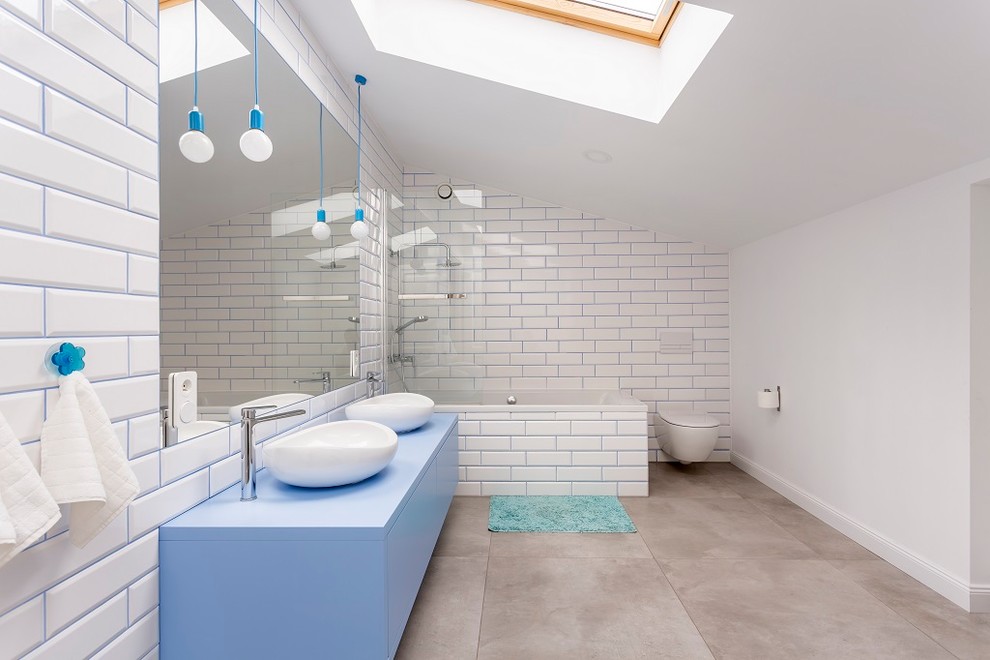 Bild på ett funkis blå blått badrum, med släta luckor, blå skåp, ett platsbyggt badkar, en dusch/badkar-kombination, en vägghängd toalettstol, vit kakel, vita väggar, ett fristående handfat och grått golv