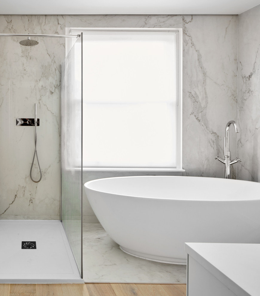 Modern inredning av ett mellanstort vit vitt badrum för barn, med vita skåp, ett fristående badkar, en öppen dusch, en vägghängd toalettstol, vit kakel, marmorkakel, vita väggar, ljust trägolv, ett integrerad handfat, brunt golv och dusch med gångjärnsdörr