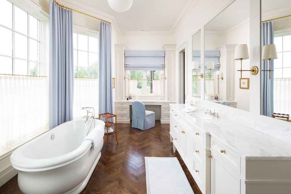 Пример оригинального дизайна: главная ванная комната в классическом стиле с белыми фасадами, отдельно стоящей ванной, белыми стенами, темным паркетным полом, врезной раковиной, коричневым полом, белой столешницей, зеркалом с подсветкой и фасадами с утопленной филенкой