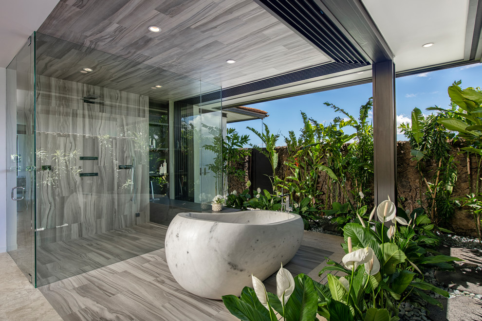 Immagine di una stanza da bagno padronale tropicale con vasca freestanding, doccia ad angolo, piastrelle grigie, pavimento grigio e porta doccia a battente
