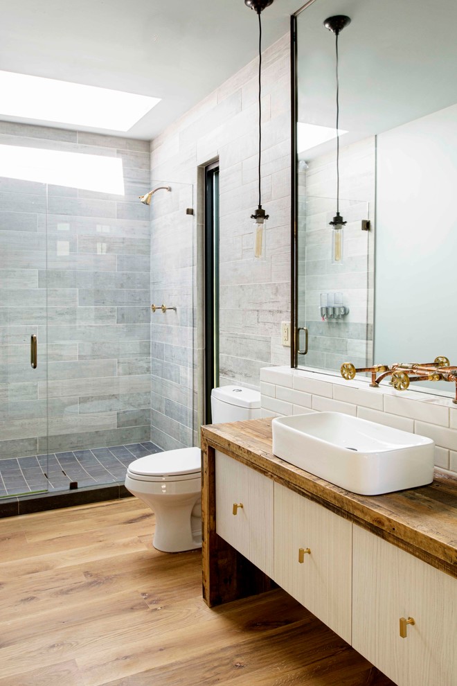 Bild på ett industriellt badrum med dusch, med släta luckor, skåp i ljust trä, grå kakel, vit kakel, ett fristående handfat, träbänkskiva, ljust trägolv och dusch med gångjärnsdörr