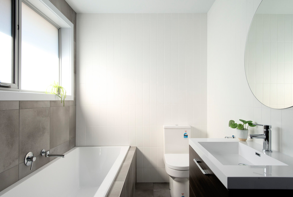 Inspiration for a small contemporary ensuite bathroom in Other with recessed-panel cabinets, dark wood cabinets, a built-in bath, a corner shower, a one-piece toilet, white tiles, ceramic tiles, grey walls, cement flooring, a wall-mounted sink, solid surface worktops, grey floors, a hinged door and white worktops.