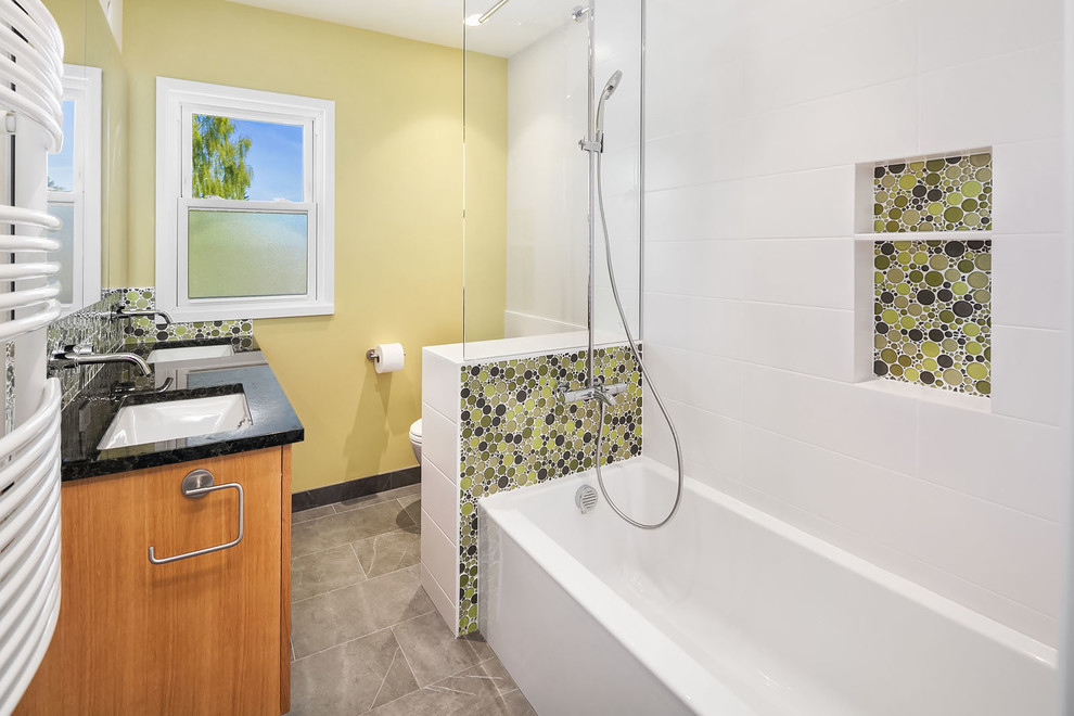 Bild på ett mellanstort shabby chic-inspirerat svart svart badrum för barn, med luckor med upphöjd panel, skåp i ljust trä, ett badkar i en alkov, en dusch/badkar-kombination, en toalettstol med hel cisternkåpa, flerfärgad kakel, glasskiva, gula väggar, klinkergolv i porslin, ett undermonterad handfat, granitbänkskiva, grått golv och dusch med duschdraperi