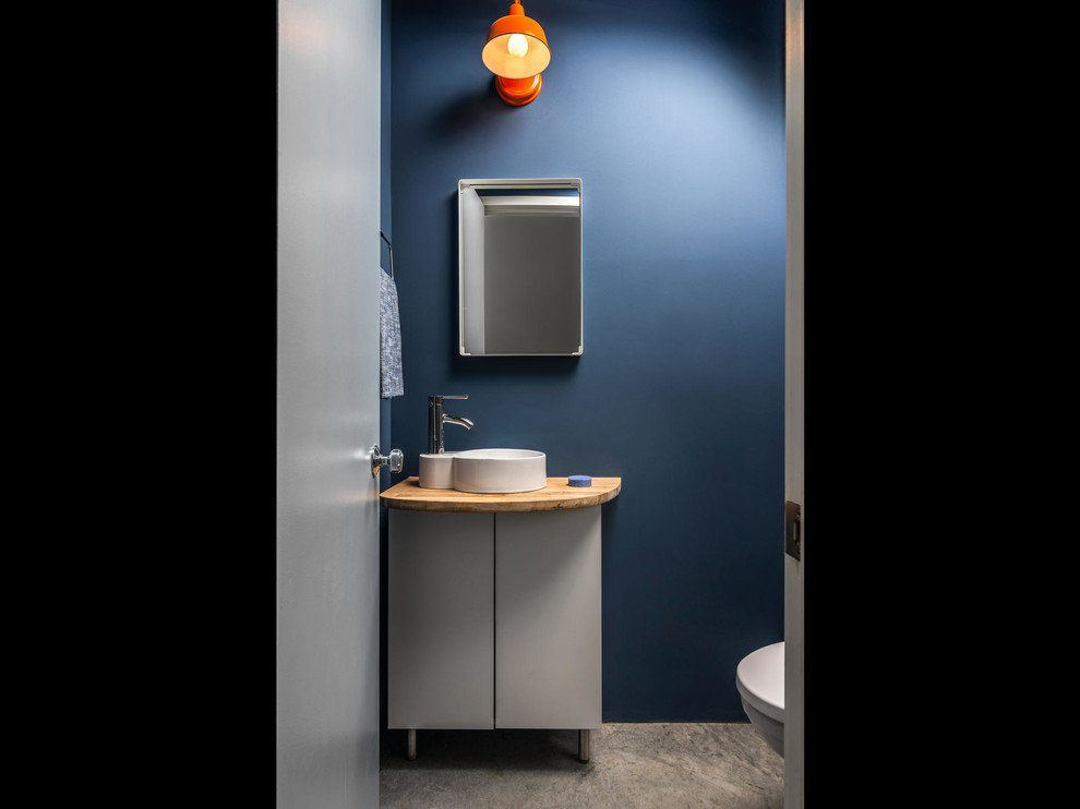 Aménagement d'une petite salle de bain industrielle avec un placard à porte plane, des portes de placard blanches, un mur bleu, sol en béton ciré, une vasque, un plan de toilette en bois et un sol gris.
