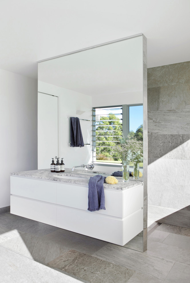 Cette image montre une salle de bain principale minimaliste de taille moyenne avec des portes de placard blanches, WC suspendus, un carrelage gris, des carreaux de porcelaine, un mur blanc, un sol en carrelage de porcelaine, un lavabo encastré, un plan de toilette en granite, un sol gris, aucune cabine, un plan de toilette gris et un placard à porte plane.