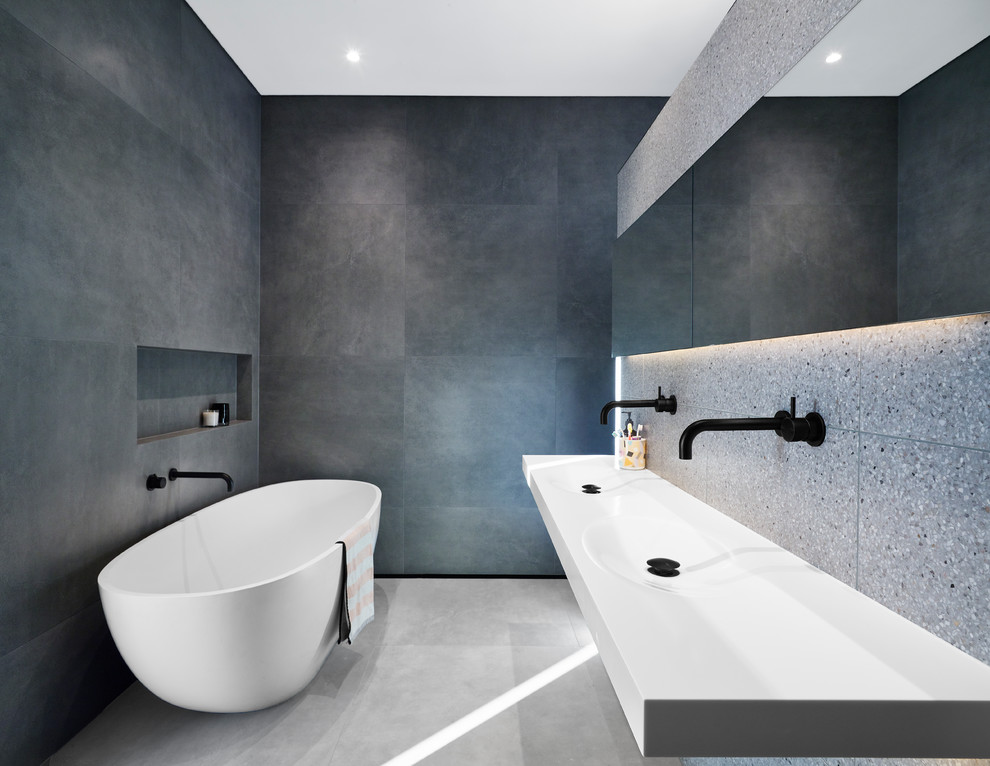 Cette image montre une salle de bain principale et grise et noire minimaliste de taille moyenne avec une baignoire indépendante, un carrelage gris, des carreaux de porcelaine, un sol en carrelage de porcelaine, un plan de toilette en surface solide, un sol gris, un lavabo intégré, un plan de toilette blanc et une niche.