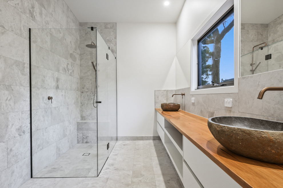 Foto på ett mellanstort funkis brun badrum med dusch, med släta luckor, vita skåp, en hörndusch, grå kakel, kakelplattor, grå väggar, kalkstensgolv, ett fristående handfat, träbänkskiva, grått golv och dusch med gångjärnsdörr