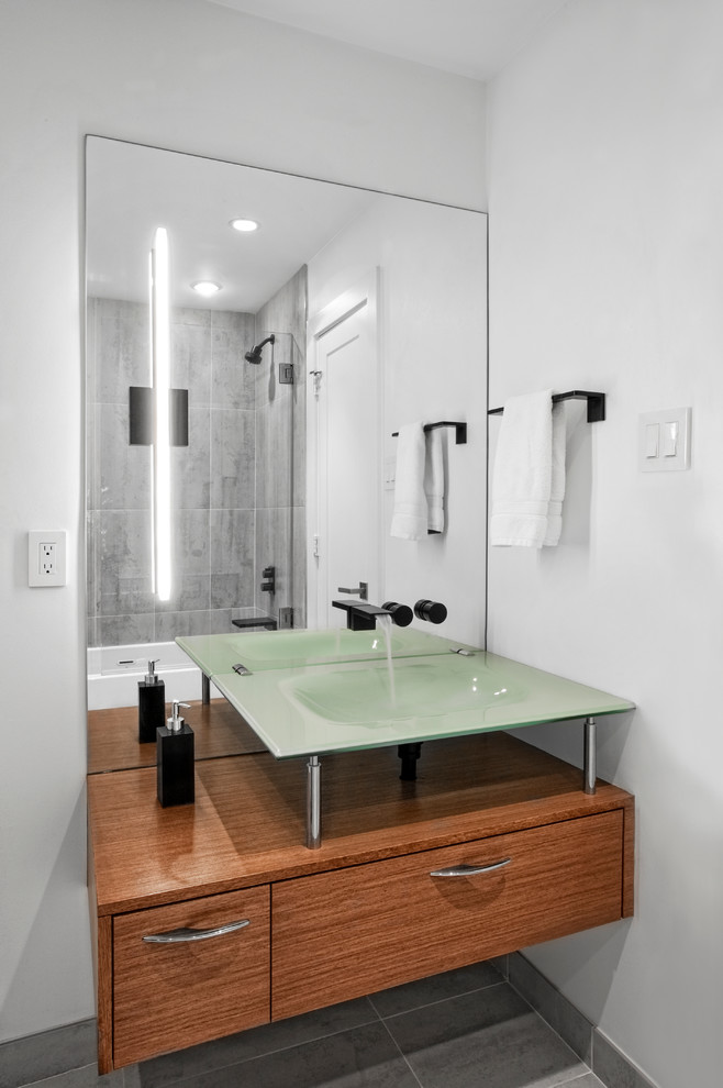 Idéer för att renovera ett mellanstort 60 tals badrum med dusch, med släta luckor, skåp i mellenmörkt trä, ett badkar i en alkov, en dusch/badkar-kombination, grå kakel, porslinskakel, grå väggar, klinkergolv i porslin, ett integrerad handfat, bänkskiva i glas, grått golv och med dusch som är öppen